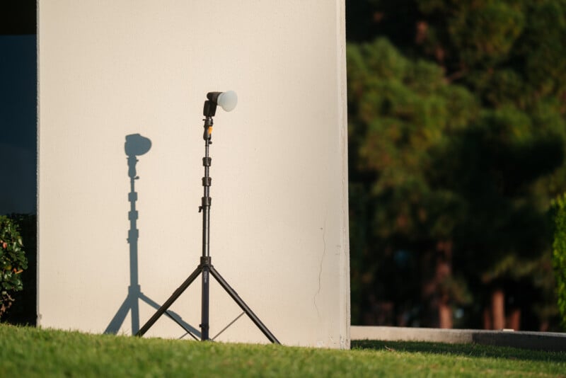 El soporte de luz fotográfica con flash proyecta sombras en una pared beige al aire libre con hierba verde en primer plano y árboles borrosos en el fondo.