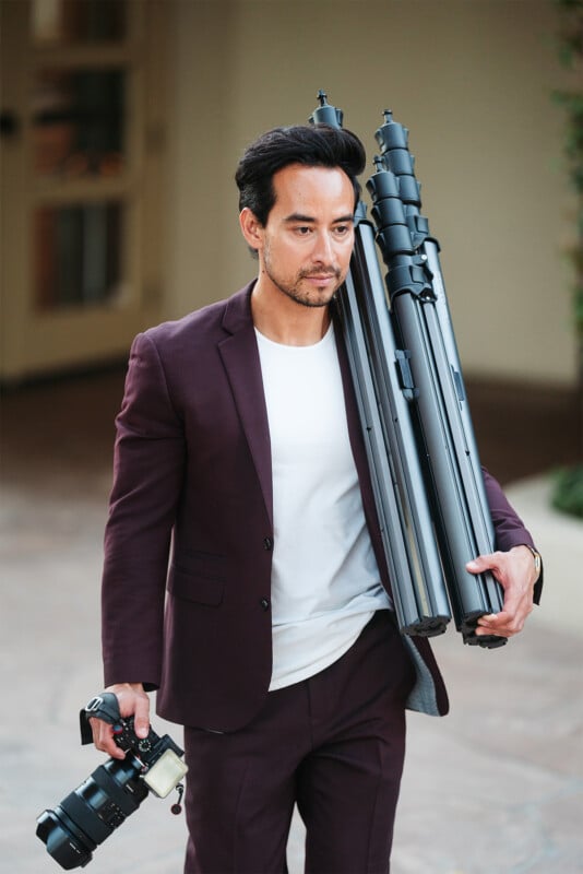 Un hombre vestido con un traje granate y una camisa blanca camina al aire libre con dos trípodes doblados sobre sus hombros y una cámara en la otra mano.