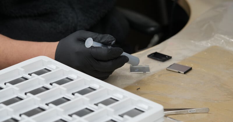 A person wearing black gloves uses a syringe-like tool near small rectangular components on a table, with a white plastic tray and tweezers nearby.