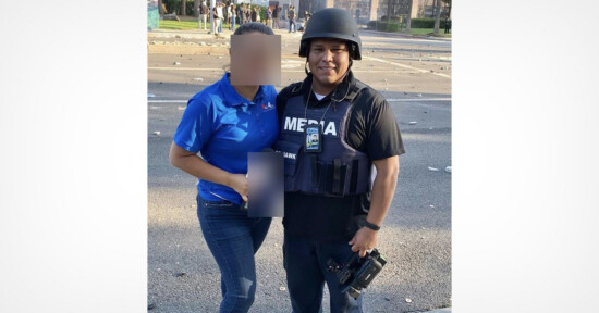 A woman in a blue shirt stands next to a man wearing a helmet, a "MEDIA" vest, and holding a camera. Their faces are blurred. Debris is scattered on the street in the background.