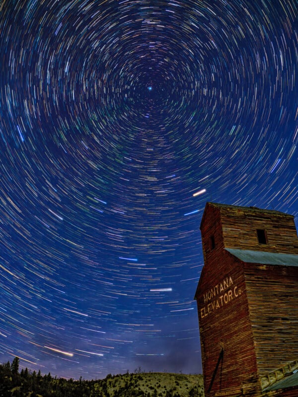 Star trails swirl around the night sky above a wooden building labeled “Montana Elevator Co.” in a rural landscape, creating a vivid circular pattern of colorful light streaks.