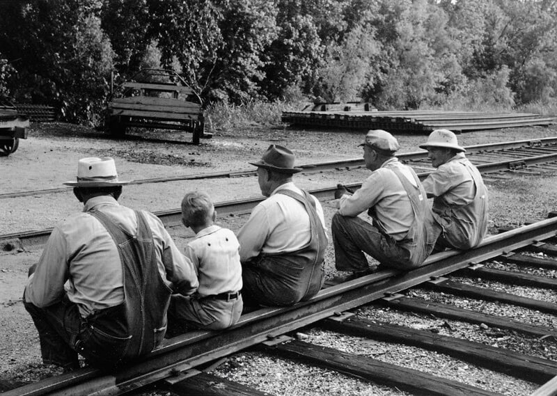 Five men and one boy wearing hats and overalls sit on a railroad track, facing away from the camera. Trees, a dirt road, and parked vehicles are visible in the background.