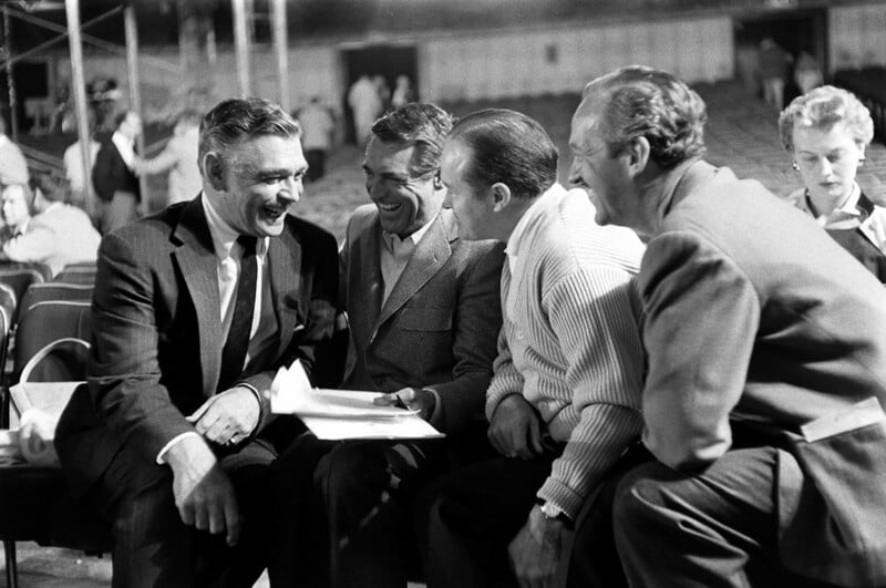 Four men in suits and jackets sit in a circle, laughing and talking together, holding papers. They are in a large, indoor space with empty chairs and other people in the blurred background.