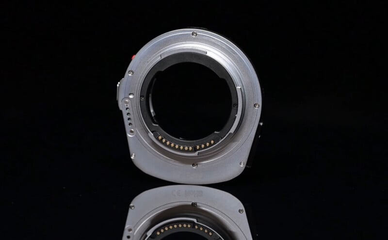 A close-up of a metallic camera lens mount adapter, standing upright against a black background with a clear reflection beneath it. The adapter features electronic contacts and several screws around its edge.
