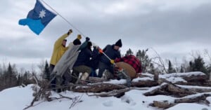Six people stand atop snow-covered logs, working together to plant a blue flag with a white star. They wear winter clothing, and a forest of bare trees is visible in the background under a cloudy sky.
