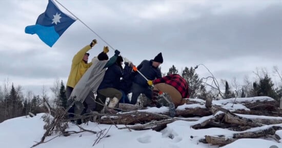 Six people stand atop snow-covered logs, working together to plant a blue flag with a white star. They wear winter clothing, and a forest of bare trees is visible in the background under a cloudy sky.