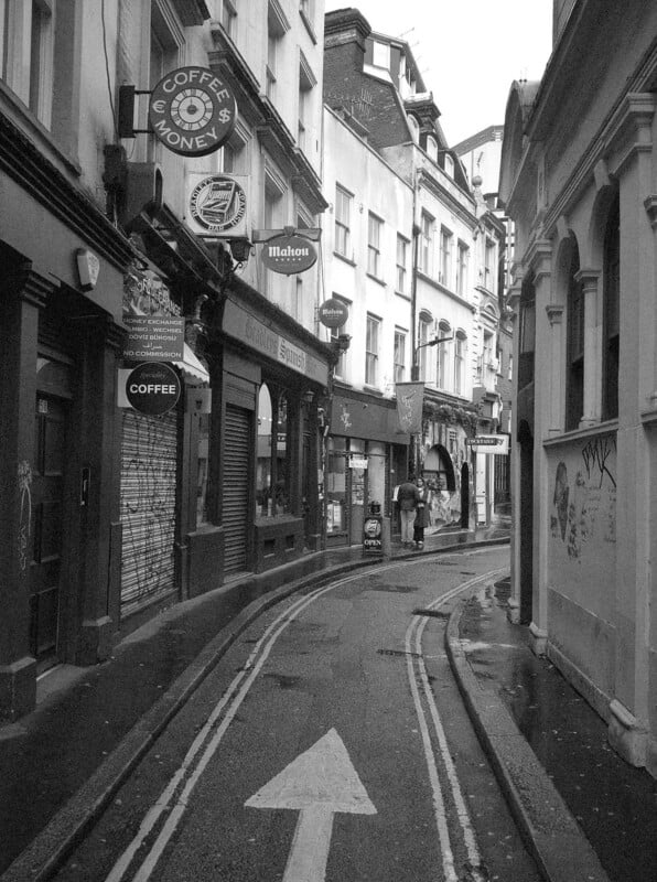 A narrow, winding street in a city, lined with old buildings and various shop signs. The road has a painted arrow and double yellow lines. Two people walk in the distance under overcast skies. The image is in black and white.