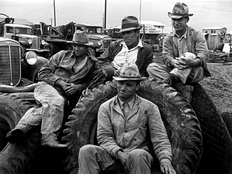 Four men in work clothes and hats sit and lean on large tractor tires in a dusty outdoor setting with trucks and machinery in the background, appearing tired and contemplative.