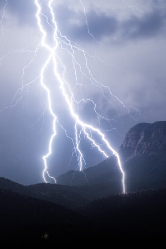 Bright lightning bolts strike the ground near dark, forested mountains under a cloudy night sky, illuminating the rugged landscape.