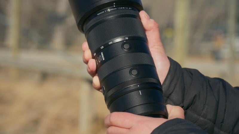 A person wearing a dark jacket holds a large black telephoto camera lens outdoors, with both hands supporting the lens; the background is blurred and sunlit.