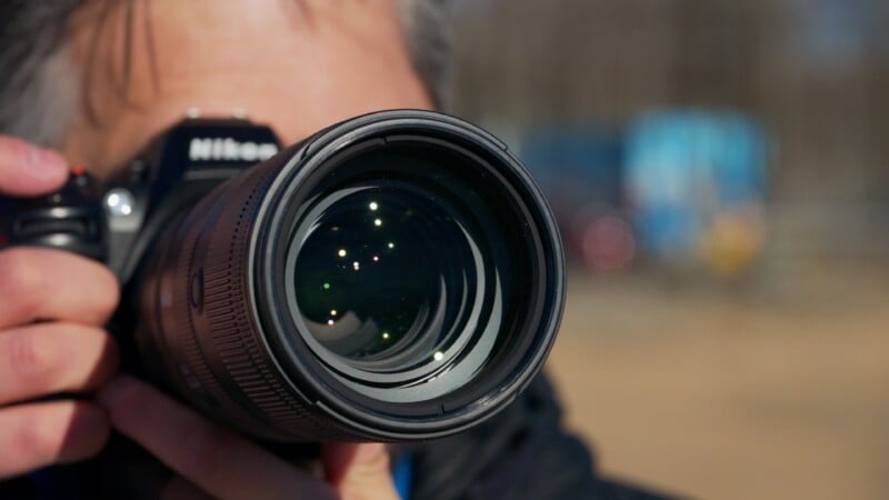 A close-up of a person holding a Nikon camera, with the focus on the large camera lens. The person's face is partially visible behind the camera. The background is blurred, suggesting an outdoor setting.