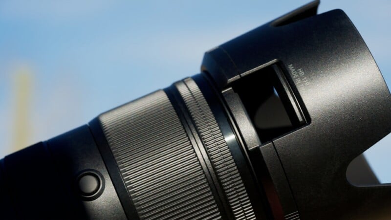 Close-up of a black camera lens set against a clear blue sky, showing detailed texture, ridges, and buttons on the lens barrel.