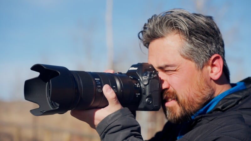 A man with gray hair and a beard is outdoors, holding a camera with a large lens up to his eye, appearing to take a photo on a sunny day.