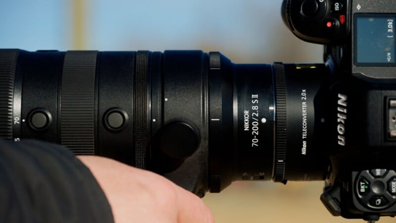 Close-up of a person’s hand holding a Nikon camera with a NIKKOR 70-200mm f/2.8 S lens and a Nikon Teleconverter 2.0x attached. The camera and lens details are clearly visible.