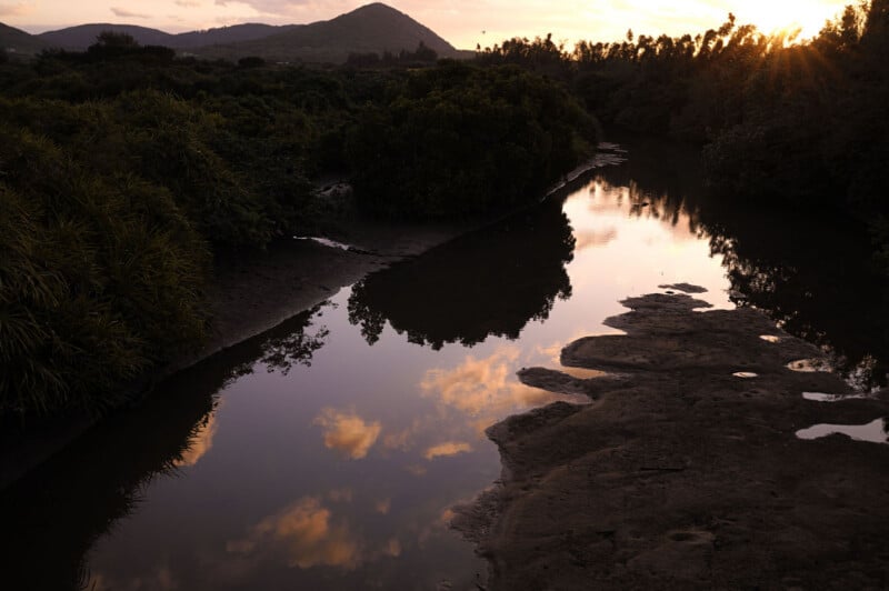A calm river winds through dense green bushes and trees at sunset, reflecting clouds and warm light in the water. Distant hills are silhouetted against an orange sky.