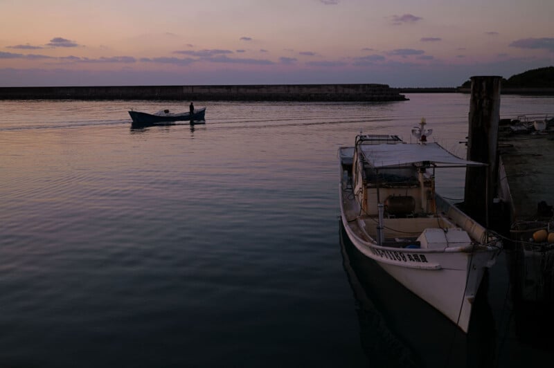 A docked white boat in the foreground and a small boat on calm water at sunset, with a breakwater in the distance and a pastel-colored sky.