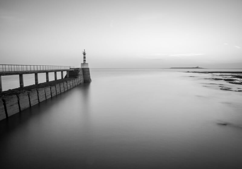 A black and white photo of a calm seascape with a long pier ending in a small lighthouse, stretching into still water. The horizon is visible with a distant island and lighthouse under a clear sky.