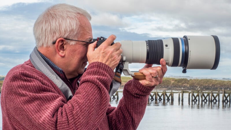 An older man with gray hair and glasses holds a large camera with a telephoto lens, aiming it towards the water with a wooden pier and cloudy sky in the background.
