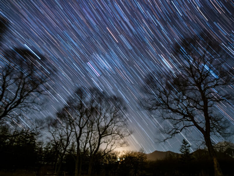 Long-exposure photo of the night sky showing star trails above silhouetted trees, with the faint glow of the moon or sun near the horizon and mountains in the background.