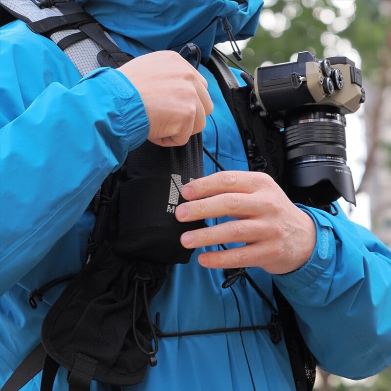 A person in a blue jacket adjusts a strap on their black chest harness while carrying a camera around their neck. The scene suggests they are preparing for outdoor photography.