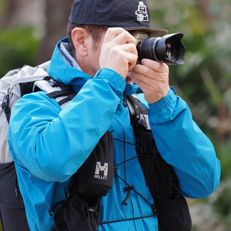 A person wearing a blue jacket and black cap is outdoors, holding a camera up to their face and taking a photo. They have a backpack and camera gear, with greenery blurred in the background.
