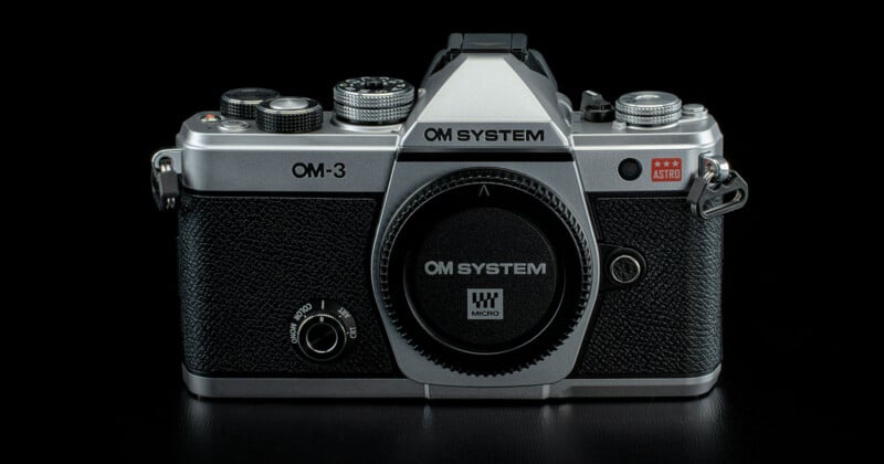 A classic OM System OM-3 film camera with a silver and black body, shown from the front against a black background. The lens mount is uncovered, and the camera branding is clearly visible.