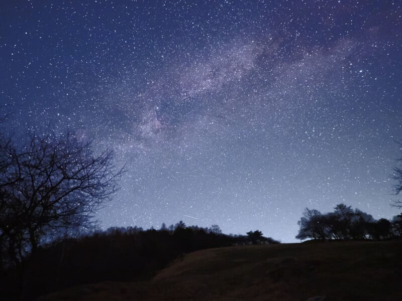 A night sky filled with stars and a visible section of the Milky Way, with silhouettes of trees and a hill in the foreground.