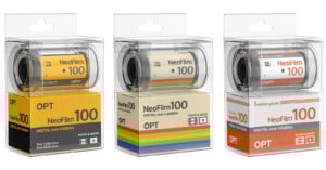 Three boxes of NeoFilm 100 digital mini cameras are displayed, each resembling a roll of film. The boxes have different colored bands: yellow, rainbow, and orange. The packaging highlights “DIGITAL mini CAMERA” and “OPT.”.