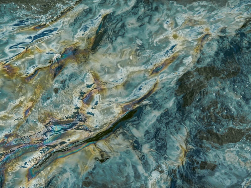 Close-up of water with swirling patterns and a visible oily sheen on the surface, creating rainbow-like colors and abstract reflections.