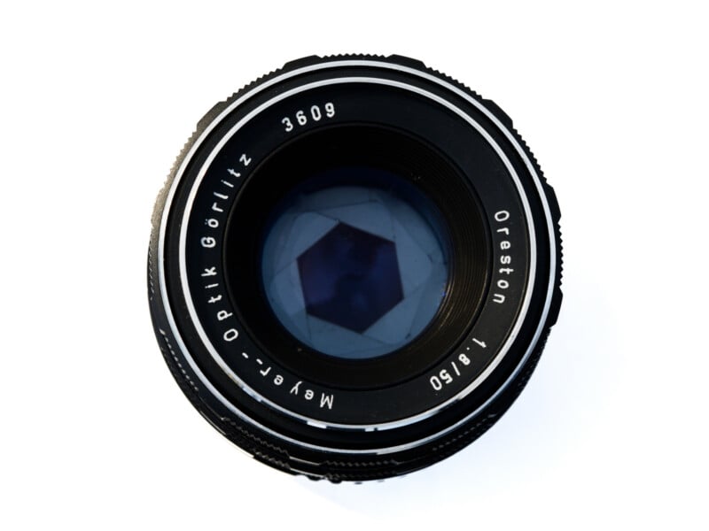 Vista cercana de la lente de la cámara Meyer-Optik Görlitz Oreston de 50 mm f/1.8 que muestra elementos de vidrio y láminas de apertura, tomada sobre fondo blanco.