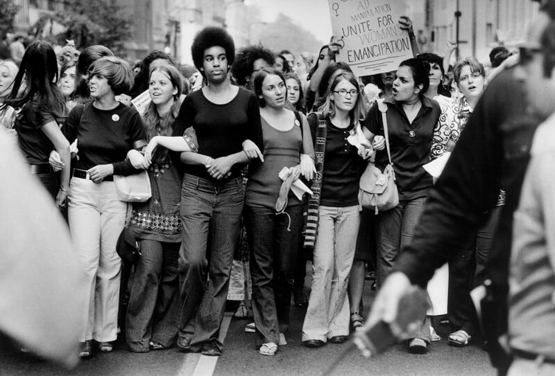 A group of women march arm-in-arm at a protest, some holding a sign that reads "Unite for Women’s Emancipation." They appear determined and united, with others marching behind them on a city street. Black and white photo.