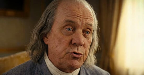 An older man with long gray hair and light skin, dressed in historical clothing with a white cravat, is sitting indoors and speaking, with a curtain and soft light in the background.