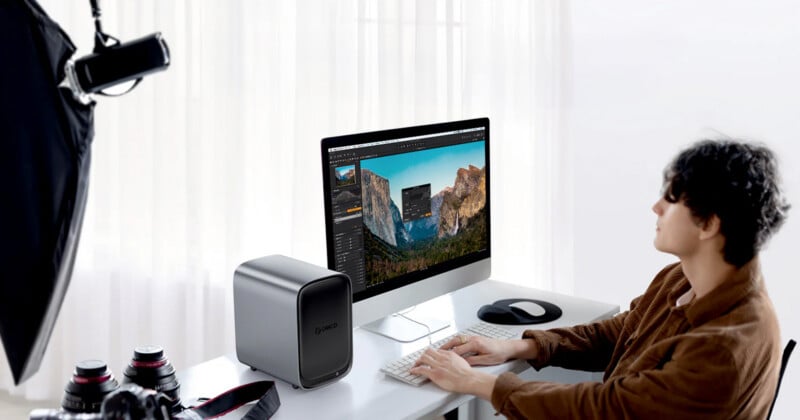 A person sits at a desk editing a landscape photo on a large desktop computer, surrounded by camera lenses, a desktop device, and studio lighting equipment in a bright, modern room.