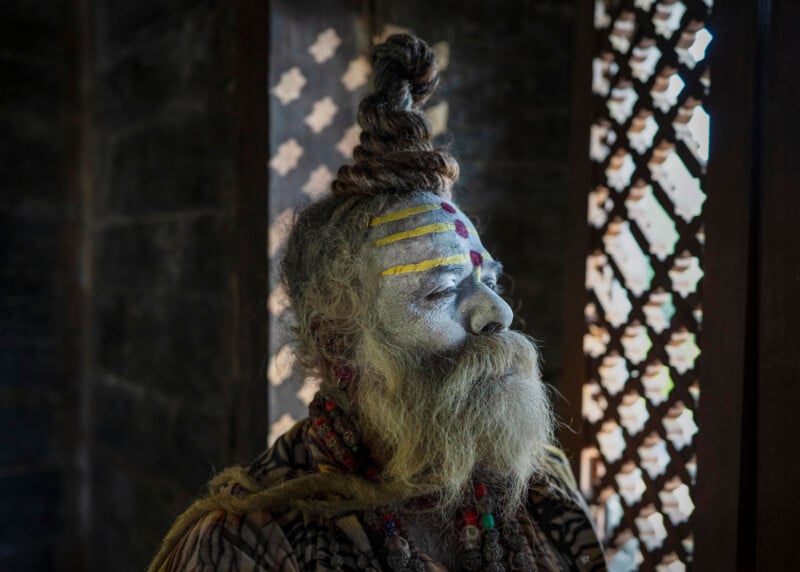 An elderly man with a long beard and hair tied in a topknot sits indoors. His face and body are covered in white ash with yellow and red paint markings on his forehead. Ornate wooden panels with geometric patterns are in the background.