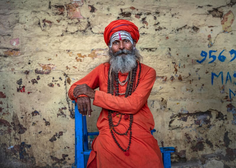 An elderly man with a long gray beard wears orange robes, a matching turban, and multiple beaded necklaces, sitting on a blue chair against a weathered, textured wall with faded markings.