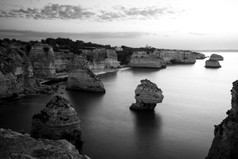 Black and white photo of dramatic coastal cliffs and large, jagged rock formations rising from calm water, with a partly cloudy sky overhead and a distant shoreline visible on the horizon.