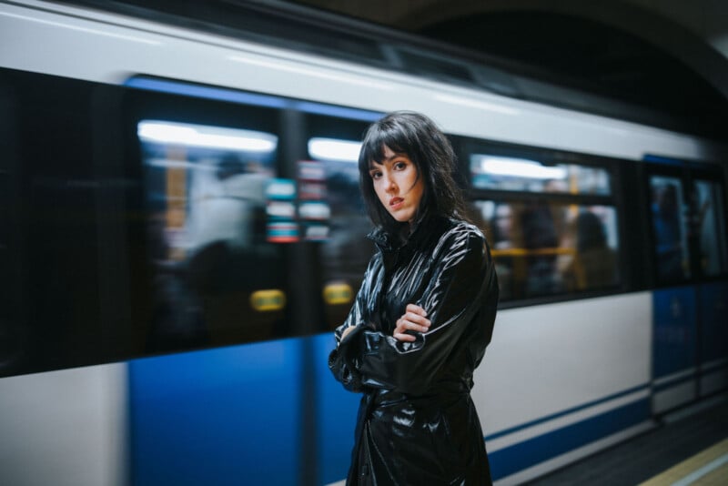 A woman in a black coat stands with arms crossed on a subway platform as a train speeds by in the background, creating a motion blur. She looks directly at the camera with a serious expression.