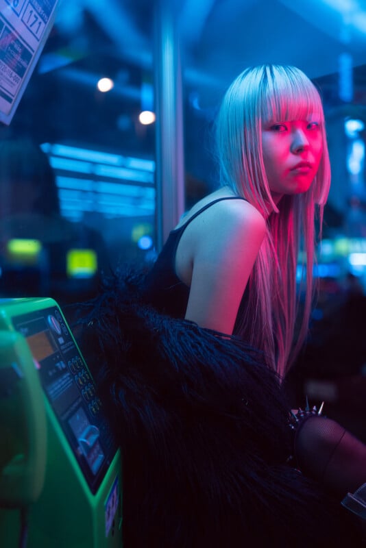 A woman with long, straight platinum blonde hair sits next to a green public payphone, bathed in neon pink and blue light, wearing a black fur coat and looking over her shoulder with a serious expression.