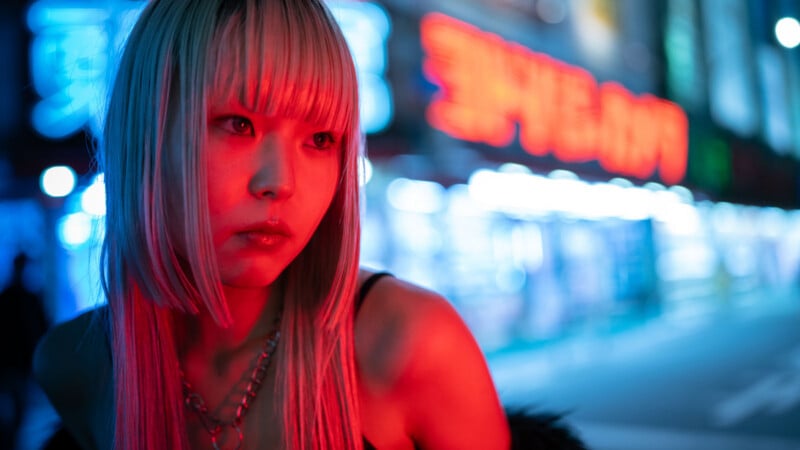 A young woman with long, straight blonde hair and bangs stands outside at night, illuminated by red and blue neon lights. She looks to the side with a serious expression, and a city street is blurred in the background.