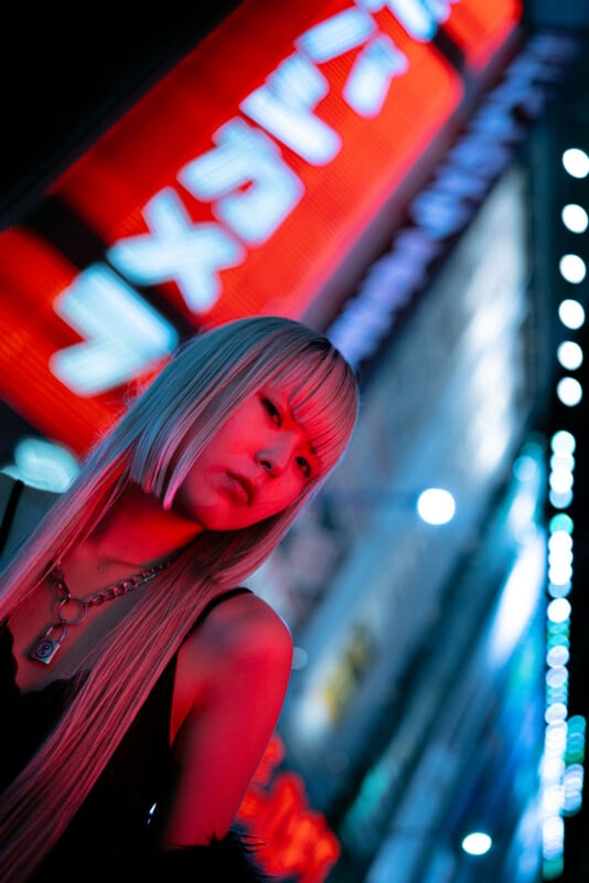 A person with long blond hair and bangs stands in a city at night, illuminated by red and blue neon lights with Japanese signs glowing brightly in the background.