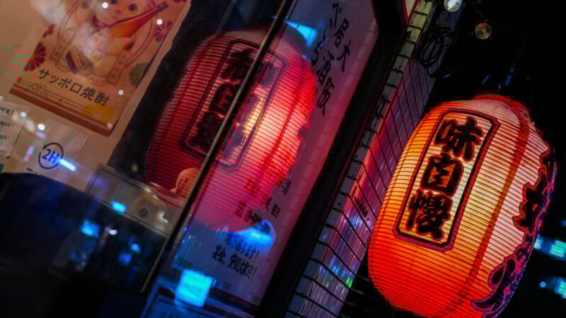 A glowing red Japanese lantern with black writing hangs outside, reflected in a window. The scene includes tiled walls, dim lighting, and Japanese posters, creating a cozy and urban nighttime atmosphere.