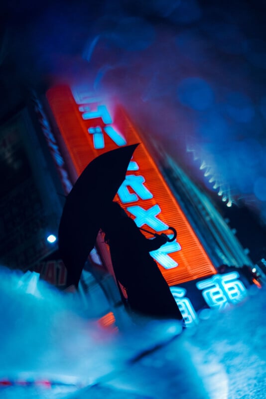 A person holding an umbrella stands in the rain at night, silhouetted against bright neon red and blue signs in a city street. The image has a moody, cinematic atmosphere with blurred reflections on the ground.