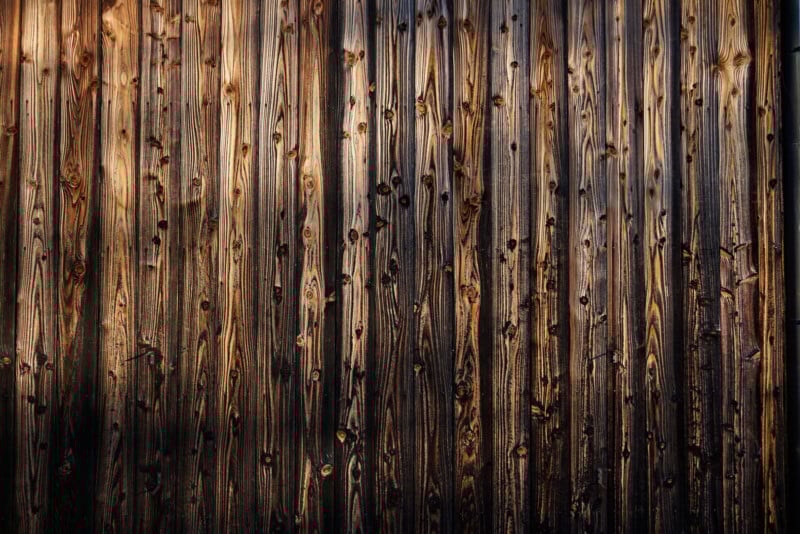 Weathered wooden planks with visible knots and grain patterns, arranged vertically. The wood has a dark, rustic appearance with streaks of light and shadow across the surface.
