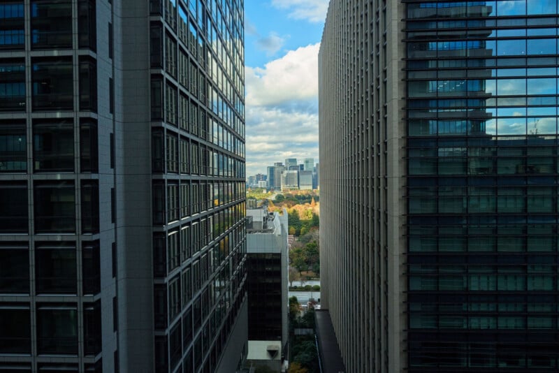 View between two tall office buildings with glass windows, looking out toward a cityscape in the distance under a partly cloudy sky. Trees with autumn colors are visible near the center of the image.