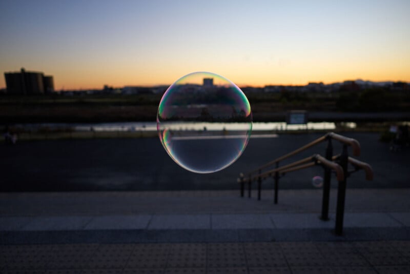 A single soap bubble floats in mid-air at sunset, with blurred steps and handrails in the foreground and a river, buildings, and an orange sky in the background.
