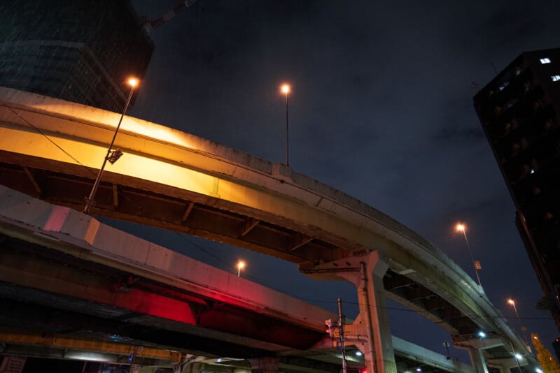 Elevated highway overpasses lit by yellow and red streetlights at night, with a dark cloudy sky and tall buildings in the background. The scene appears wet, suggesting recent or ongoing rain.