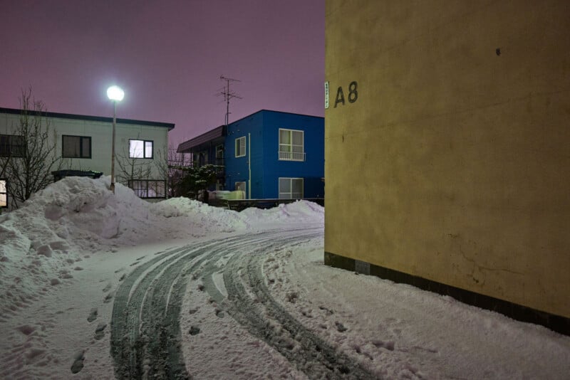 A snowy street curves past a yellow building labeled "A8" at night. Snow piles line the road, and tire and footprints are visible. A glowing streetlamp illuminates nearby houses, including a bright blue one. The sky is purple.