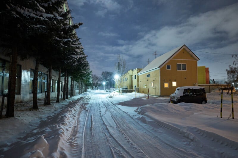 A snowy residential street at night, lined with trees and illuminated by streetlights. Tire tracks are visible in the snow, and a few houses and a parked car are seen on the right side under a partly cloudy sky.