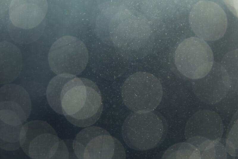 Abstract image with large, soft, out-of-focus light circles against a dark background, creating a dreamy and ethereal bokeh effect. Fine particles or dust are visible throughout the image.
