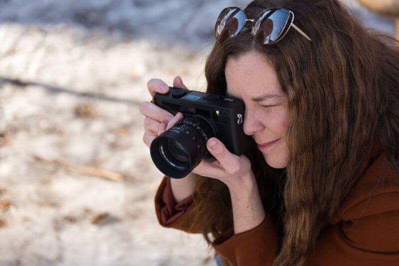 A woman with long brown hair and sunglasses on her head is outdoors, holding a camera up to her face and taking a photograph, with a blurred, sunlit background.